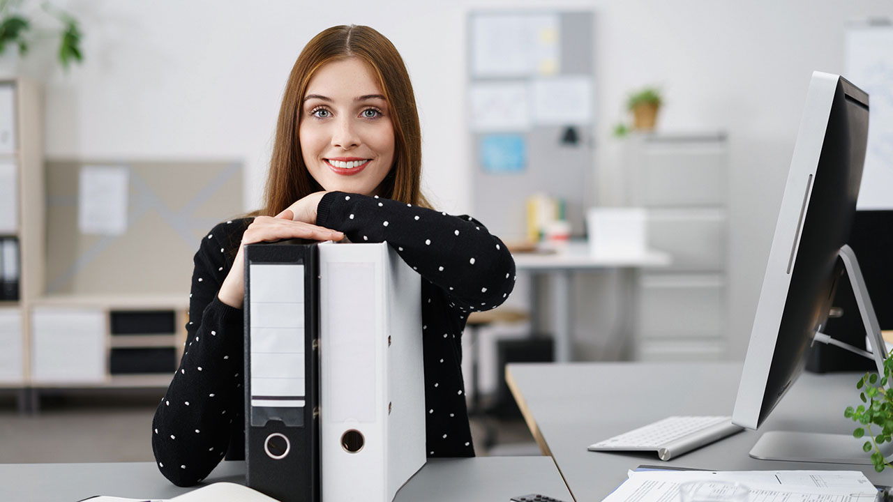 Junge lächelnde Person stützt sich mit den Armen auf zwei Aktenordnern ab, neben ihr steht ein Computer