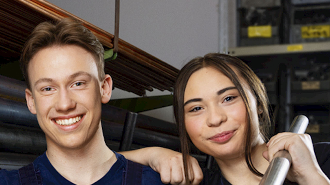 Zwei junge Personen stehen vor einer Halterung mit schwarzen Rohren und lächeln in die Kamera
