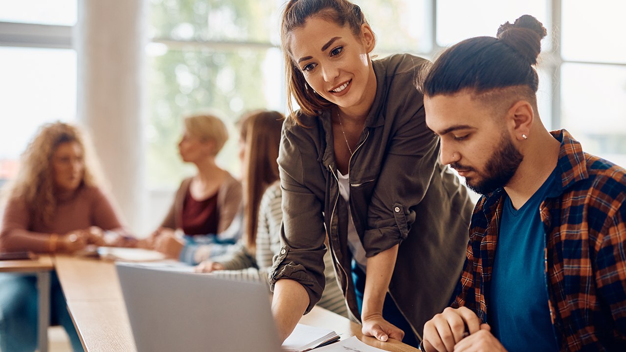 Junge Frau steht neben jungem Mann, der an einem Tisch sitzt und arbeitet. Im Hintergrund sieht man eine weitere Schülergruppe.