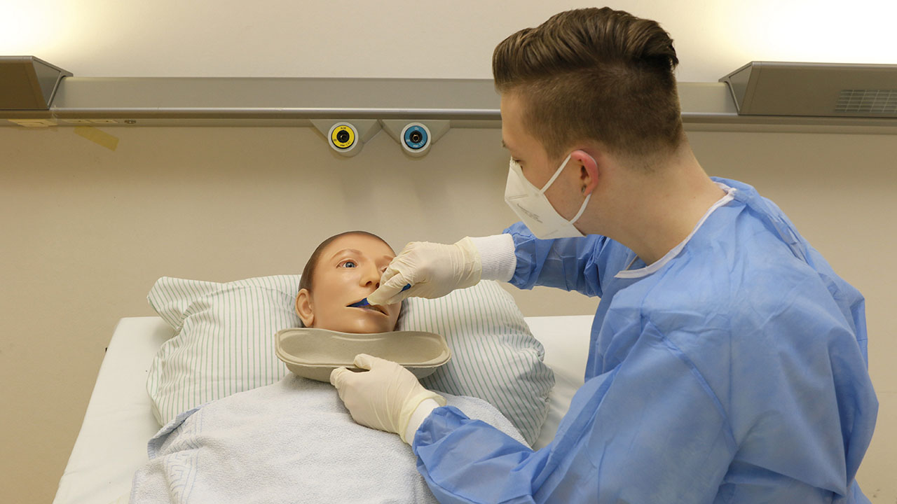 Junge Person in blauer Pflegebekleidung mit Mundschutz übt Füttern an einer Pflegepuppe