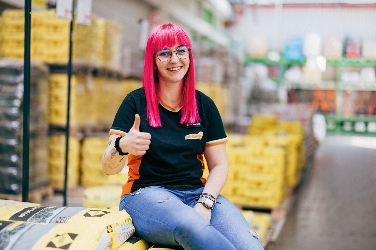 Jungendliche Azubi mit roten Haaren und Tatoos auf dem rechten Arm sitzt auf einem Stapel Säcke und zeigt den Daumen nach oben.