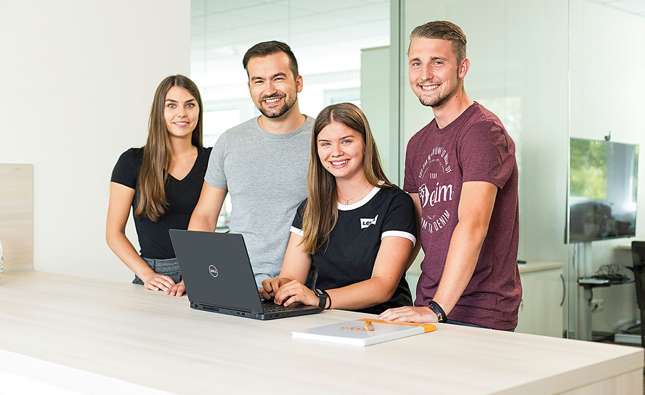 4 Junge Menschen stehen an einem Schreibtisch, auf dem ein Laptop steht.