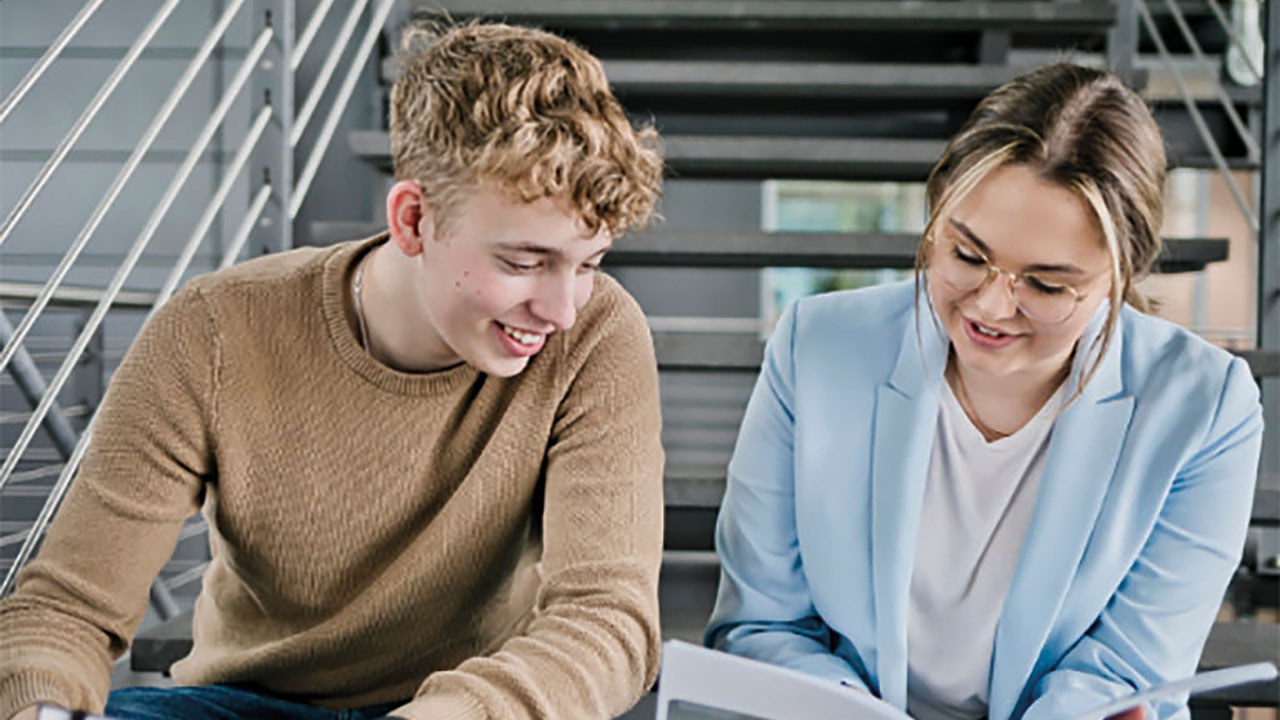 Zwei Personen sitzen auf einer Treppe und sehen gemeinsam in ein Buch, sie lächeln dabei.