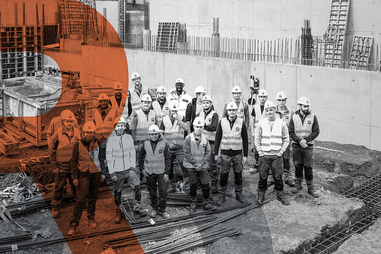 Eine größere Gruppe von Bauarbeitenden steht auf einer Baustelle zwischen Werkzeug, Stahlstangen und Schalungen. Alle tragen Schutzhelme und Warnwesten. Der Hintergrund zeigt Betonwände und Baugerüste.