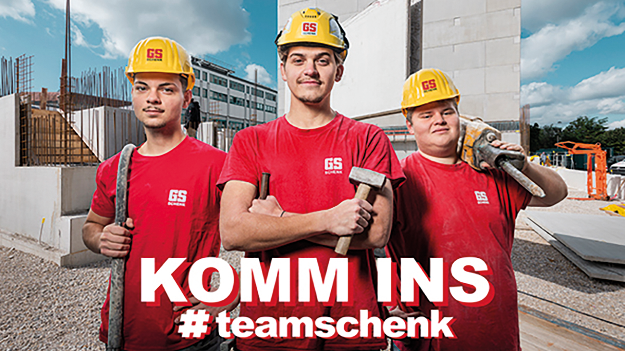 3 junge Mitarbeitende in GS Schenk-Arbeitskleidung mit gelben Helmen und roten T-shirts stehen mit Werkzeugen aus der Baubranche auf einer Baustelle. Dazu der Spruch KOMM INS #teamschenk