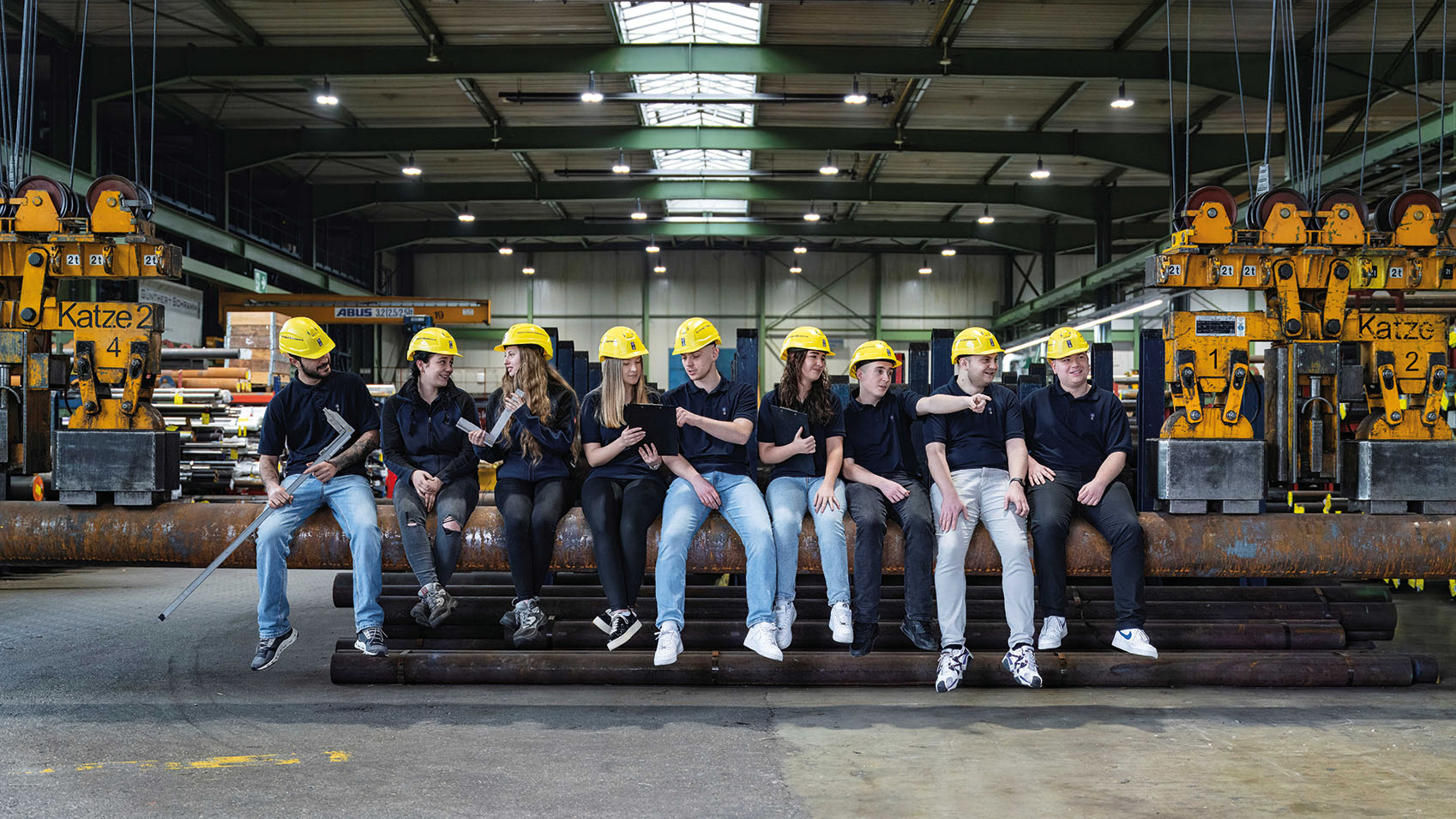 Junge Menschen sitzen in einer Lagerhalle auf einem Eisenrohr. Sie tragen gelbe Schutzhelme und haben teilweise Werkzeuge in der Hand.