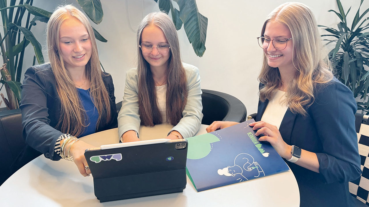 Drei junge lächelnde Personen sitzen zusammen an einem Tisch und schauen auf ein Tablet