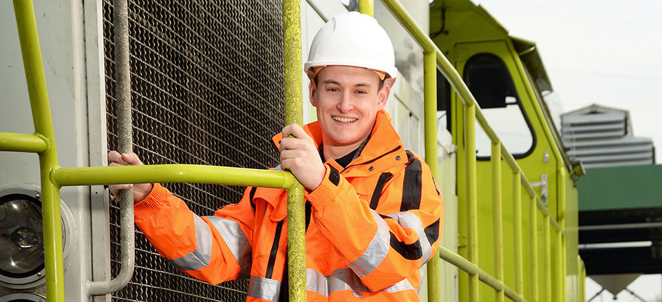 Junge lächelnde Person in orangefarbener Jacke mit Helm an einem Zug