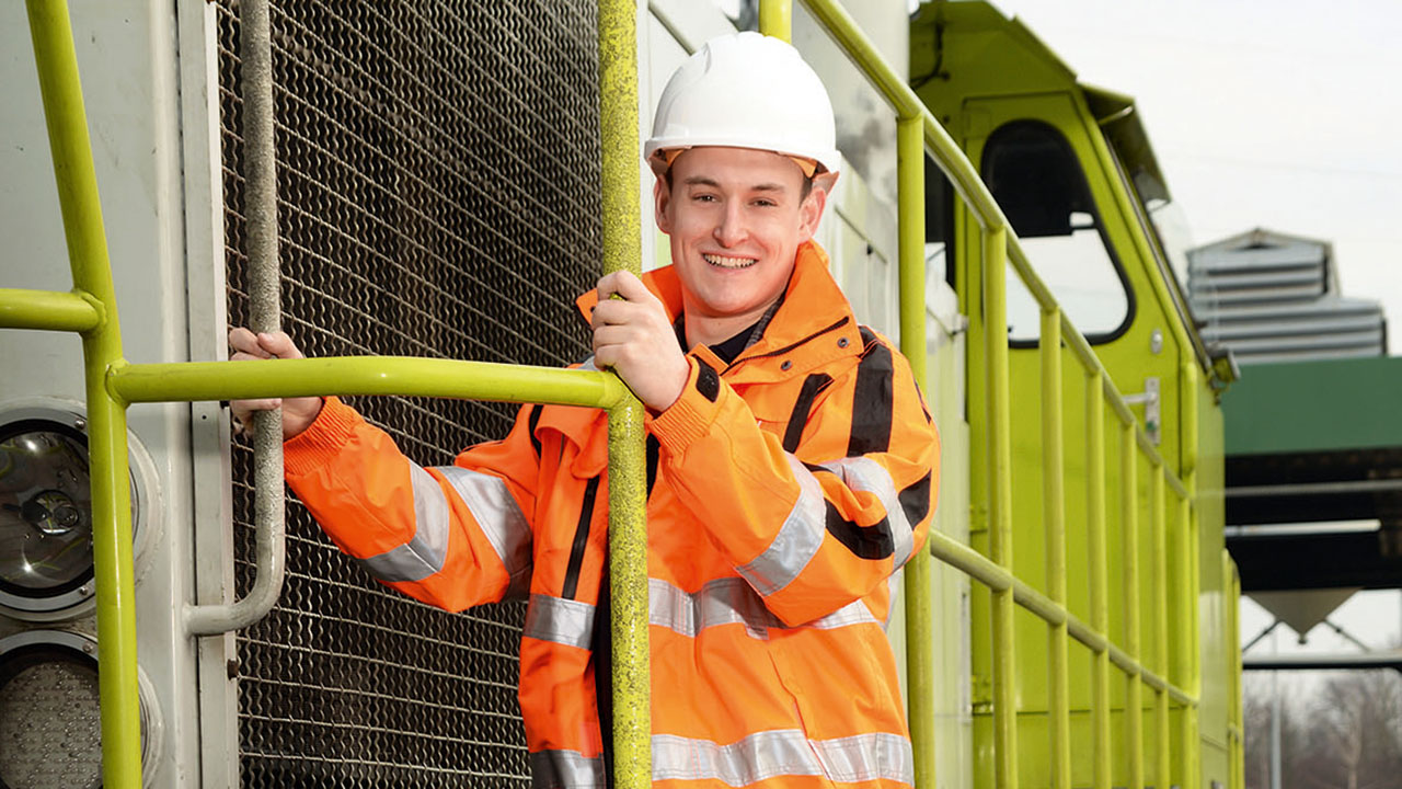 Junge lächelnde Person in orangefarbener Jacke mit Helm an einem Zug