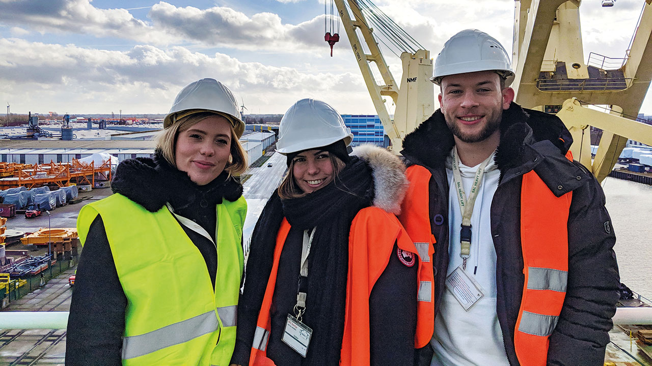 Drei junge lächelnde Personen mit Warnweste und Helm am Hafen