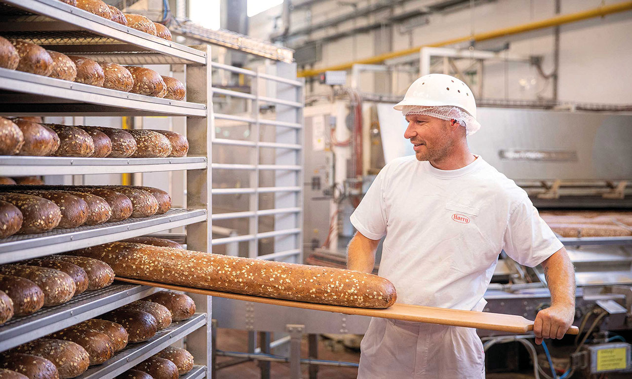 Eine Person in Bäckereikleidung schiebt mit einem Holzschieber ein langes Brot auf ein Regal voller frisch gebackener Brote. 