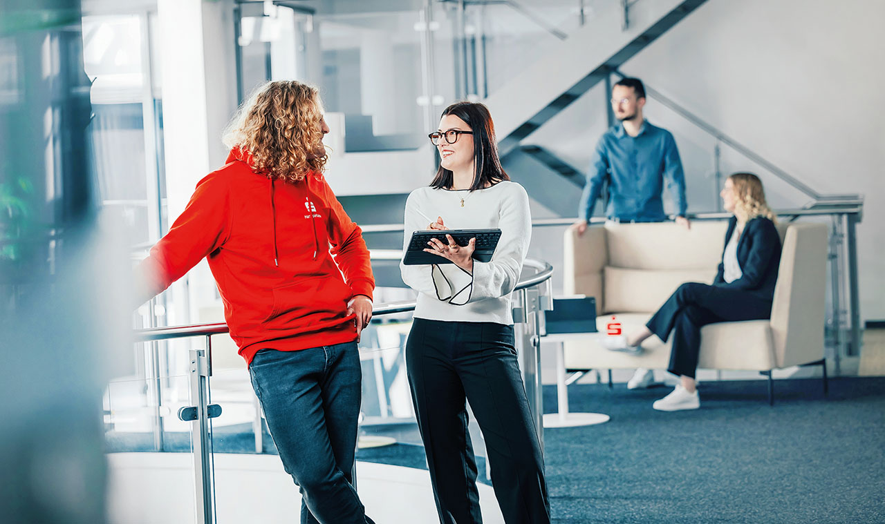 Im Vordergrund junge Dam mit braunen haaren und Brille, die einen Laptop in der Hand hält. Daneben steht ein Mann mit blonden längeren lockigen Haarn und einem roten Hoodie. Sie unterhalten sich. Im Hintergrund zwei Personen an einer Sitzecke.