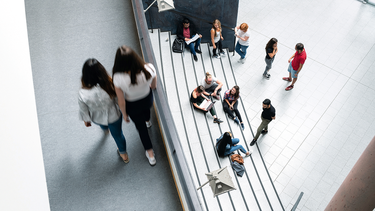 Foto in einem Gebäude der HAWK aus einer oberen Etage aufgenommen. Zu sehen sind zwei Studierende die einen Gang entlanglaufen und auf einer tieferen Ebene sitzen Studierende in kleinen Gruppen zusammen auf einer Treppe. 