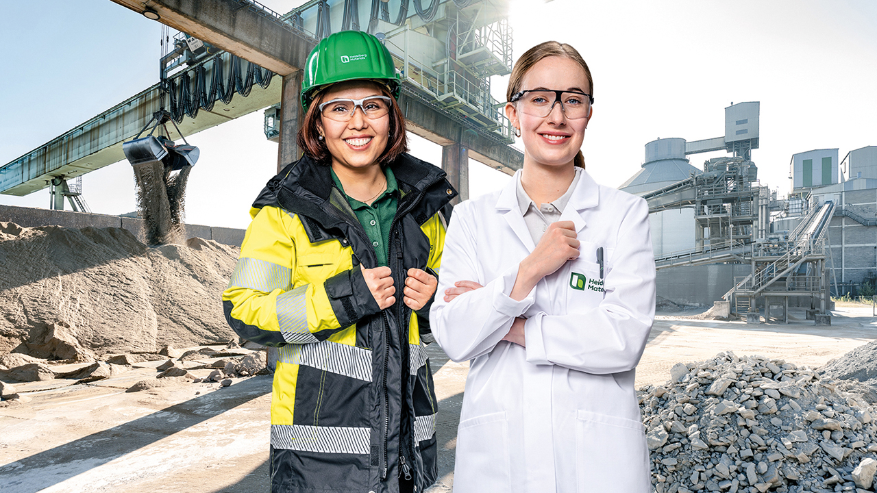 Zwei junge Frauen auf einer Baustelle, eine in Arbeitskleidung und mit Helm, die andere in einem weißen Kittel