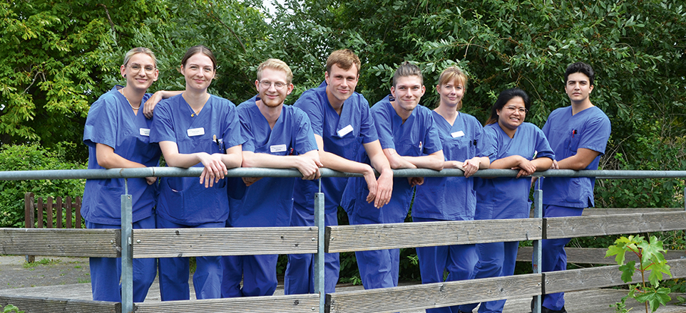 8 Mitarbeitende der Helios Klinik in ihrer blauen Dienstkleidung draußen nebeneinander an einem Geländer posierend