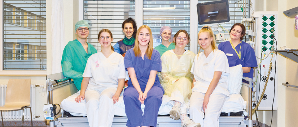 Mitarbeitende in ihrer jeweiligen Arbeitskleidung aus den verschiedenen Bereichen der Helios Klinik auf einem Krankenbett versammelt