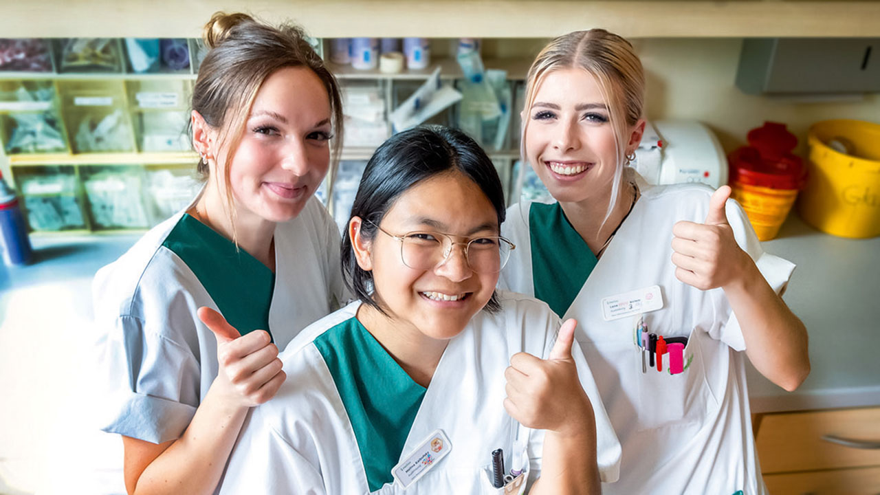 Drei junge lächelnde Personen in grün-weißer Pflegebekleidung zeigen den Daumen hoch