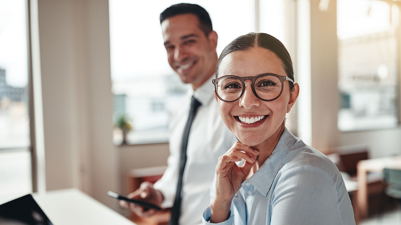 Junge Person in Businesskleidung mit Brille lächelt in die Kamera, im Hintergrund sitzt eine weitere lächelnde Person mit einem Handy in der Hand