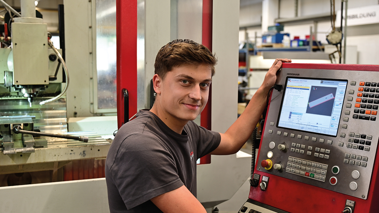 Eine Person steht an einer Maschine und lächelt in die Kamera.