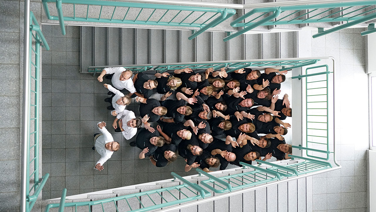 Eine Gruppe von etwa 40 Personen steht dicht beisammen in einem Treppenhaus und winkt nach oben zur Kamera. Alle tragen dunkle oder weiße Kleidung. 