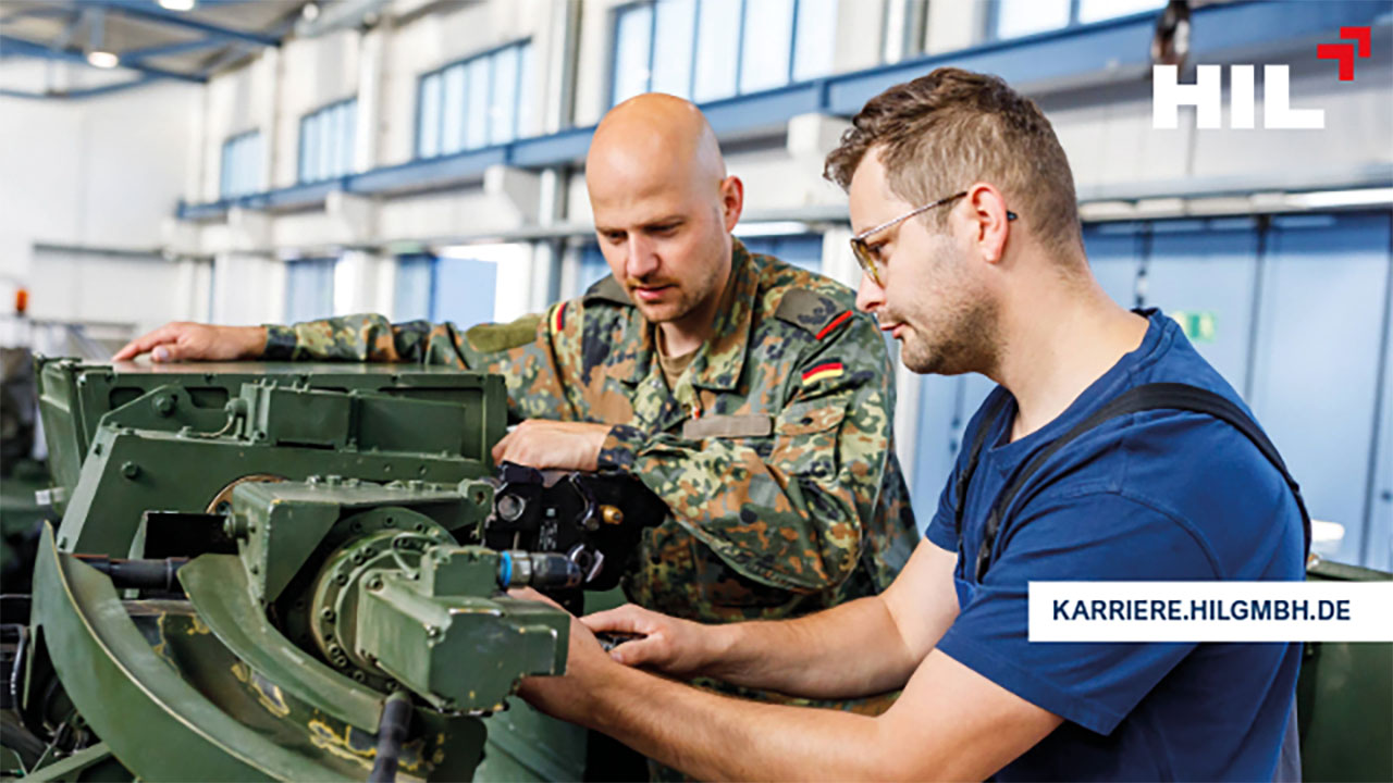 Ein Soldat und ein Mann in blauen Arbeitsanzug arbeiten zusammen an einer dunkelgrünen Maschine.