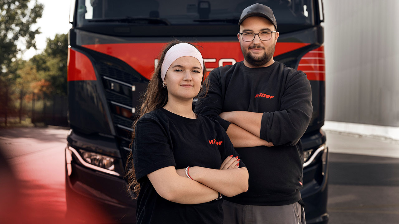 Zwei junge Menschen in schwarzen T-Shirts mit Firmenlogo stehen mit verschränkten Armen vor einem LKW
