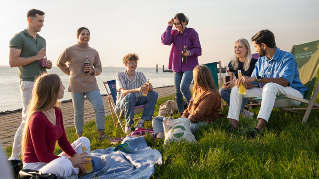 Studierende sitzen im Gras und auf Liegestühlen am Wasser und tauschen sich aus