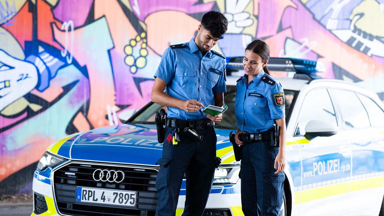 Zwei Poliizeibeamte stehen vor einem Dienstwagen. Dieser parkt vor einer Wand die mit buntem Graffiti besprüht ist.