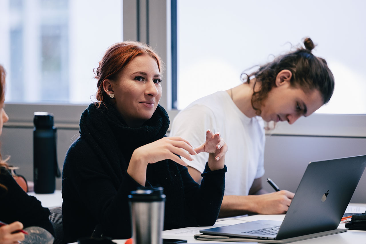 Studentin sitzt an einem Tisch und erzählt etwas. Dabei benutzt sie ihre Hände als Körpersprache.