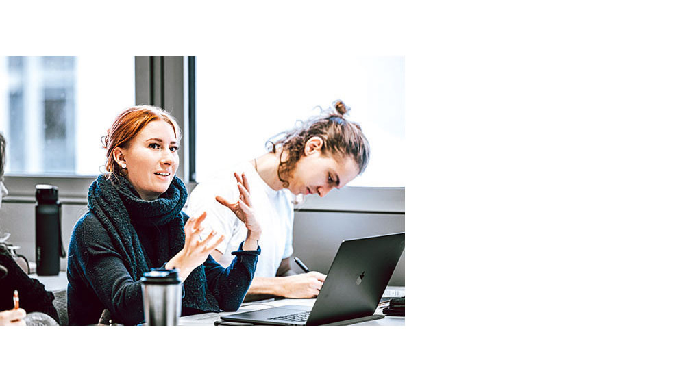 Zwei Personen sitzen an einem Tisch mit Laptop. Eine Person erklärt etwas, die andere schreibt konzentriert.