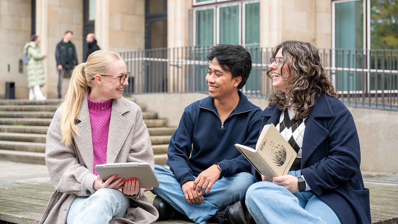 3 Studierende sitzen vor der HTW auf einer Bank und haben Buch bzw. Tablet in der Hand und unterhalten sich freudig angeregt.