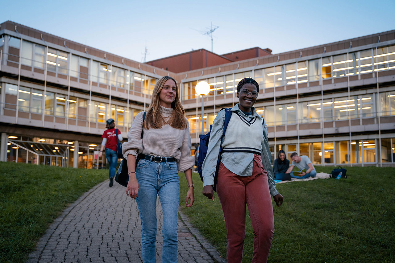 Zwei Personen gehen lächelnd einen gepflasterten Weg vor einem großen, hell erleuchteten Gebäude entlang. Im Hintergrund sitzen weitere Menschen auf dem Rasen.