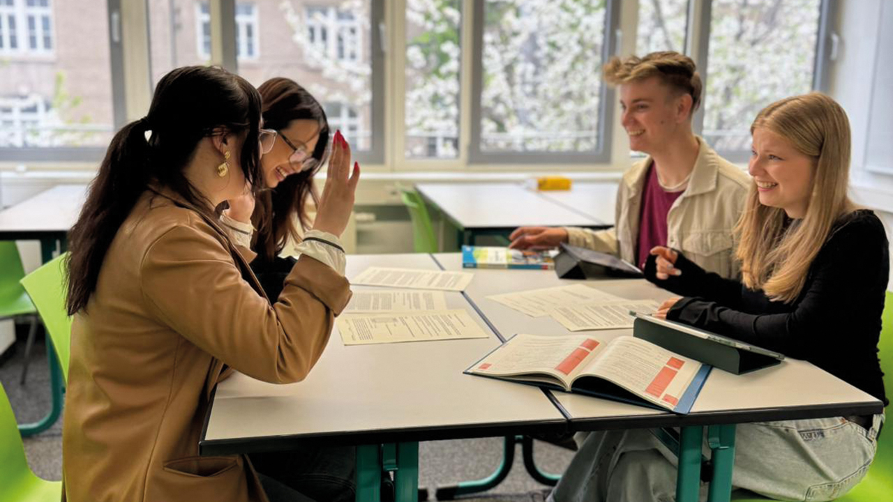 4 Schüler/innen an einem Tisch beim Arbeiten im Klassenraum. Alle sind gut gelaunt.