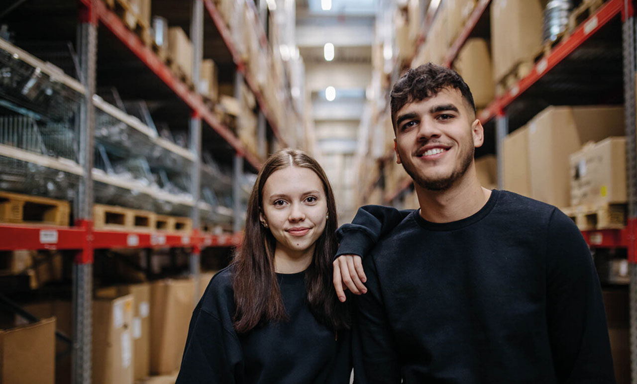 Zwei Personen in schwarzen Pullovern stehen in der Werkshalle mit vielen großen Regalen.