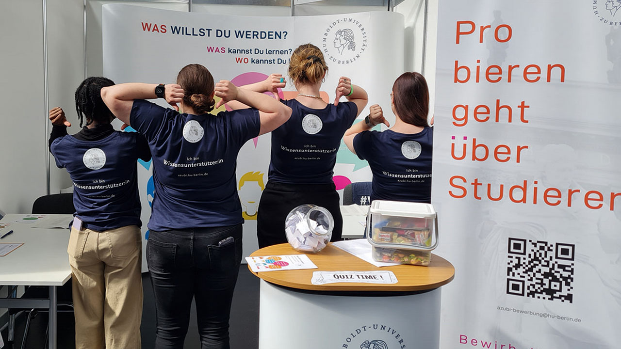 Mitarbeitende der Humboldt Universität zu Berlin am Messestand zeigen mit den Daumen auf das Logo auf dem Rücken der T-Shirts