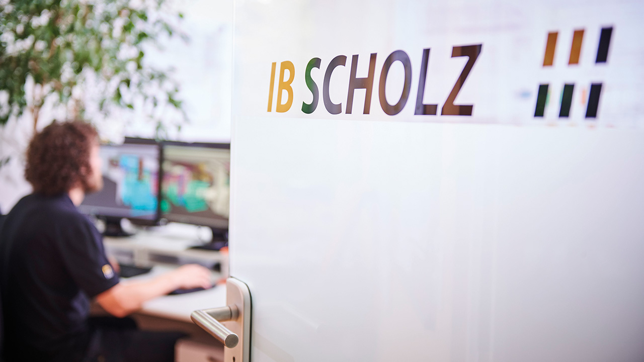 Sich öffnende Bürotür mit IB Scholz-Aufschrift zu einem Raum, wo Mitarbeitende an Schreibtisch mit Computer sitzen