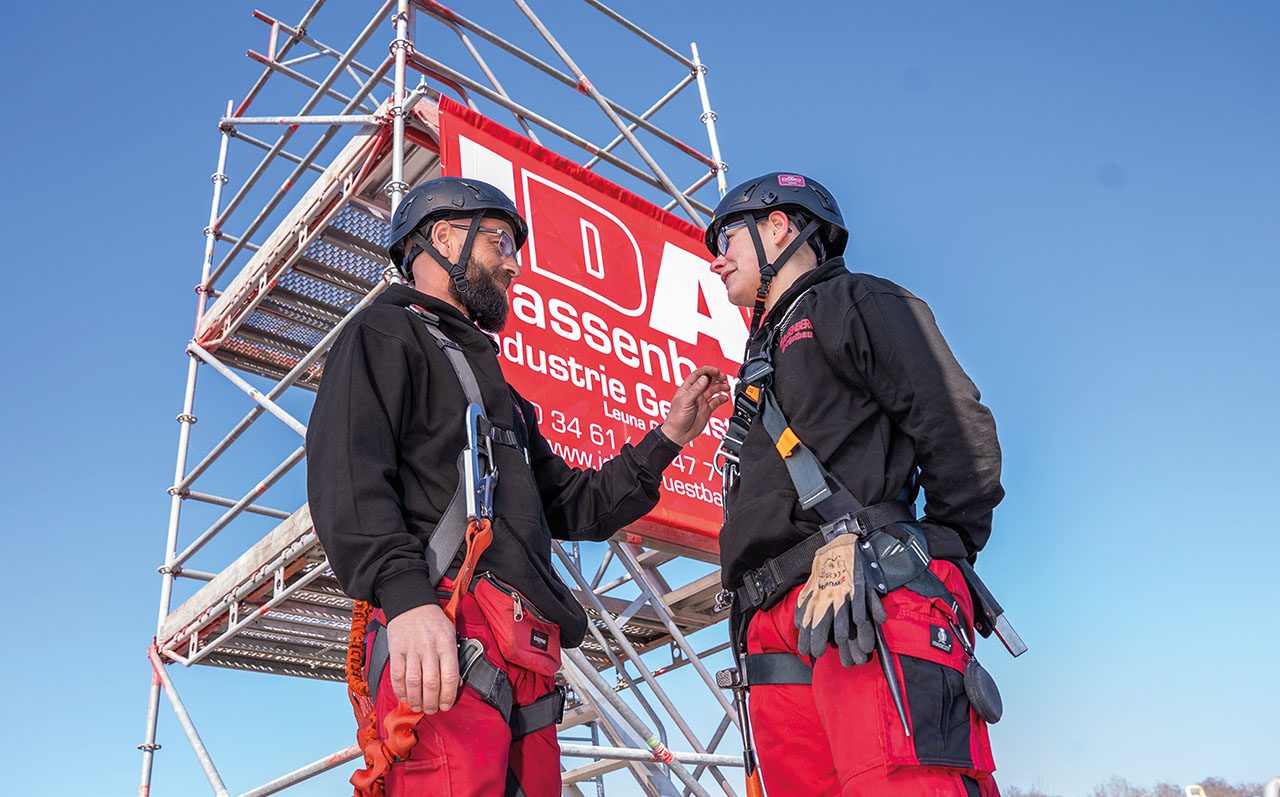 Zwei personen in rot-schwarzen Arbeitsanzügen und Helm unterhalten sich unter einem Gerpst von IDA.