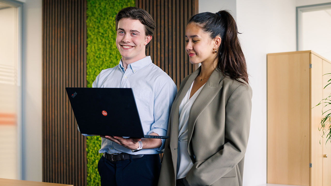 Zwei junge lächelnde Personen in Businesskleidung nebeneinander, eine Person hält einen Laptop in der Hand