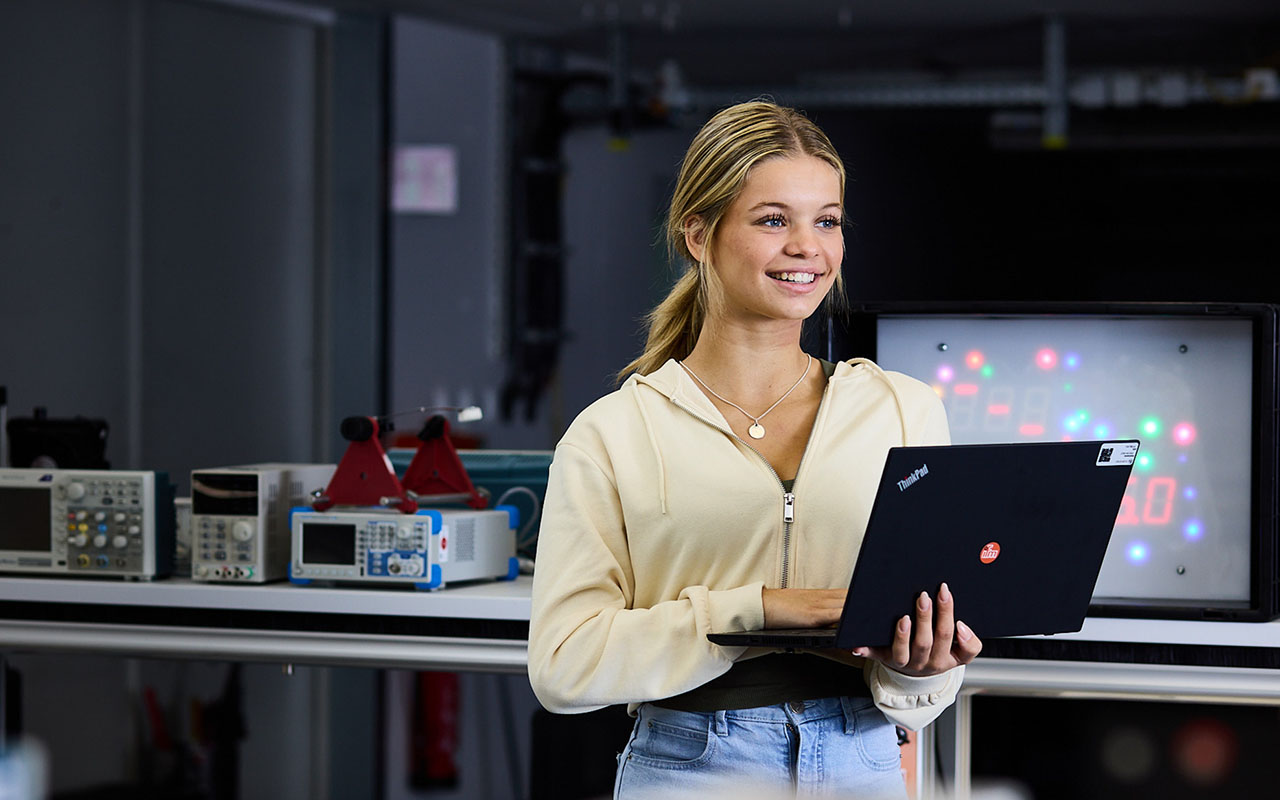 Person mit Laptop steht in einem Techniklabor und lächelt. Im Hintergrund sind Messgeräte und ein Display mit LEDs sichtbar