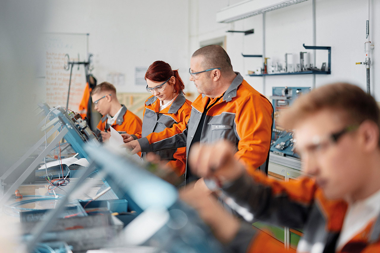 Vier Mitarbeiter in orange-grauen Arbeitsanzügen bei der Arbeit