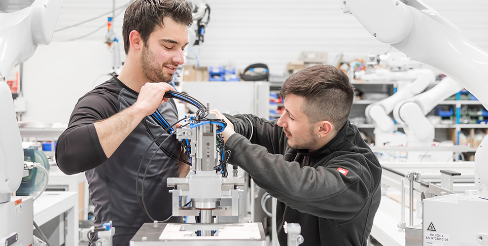 Auf dem Bild sieht man zwei junge Männer, die gemeinsam an einem technischen Aufbau arbeiten. Sie befinden sich in einer modernen industriellen Umgebung. Die Szene deutet auf eine Ausbildungs- oder Fertigungssituation hin.
