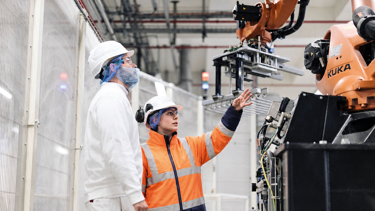 Zwei Personen in Arbeitsanzügen und Helm stehen vor einer KUKA-Maschine.