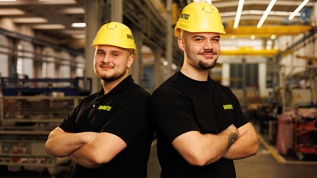 Zwei Mitarbeitende stehen mit verschränkten Armen nebeneinander in einer Werkshalle von Innomotics. Sie tragen gelbe Helme und lächeln den Betrachtenden an.