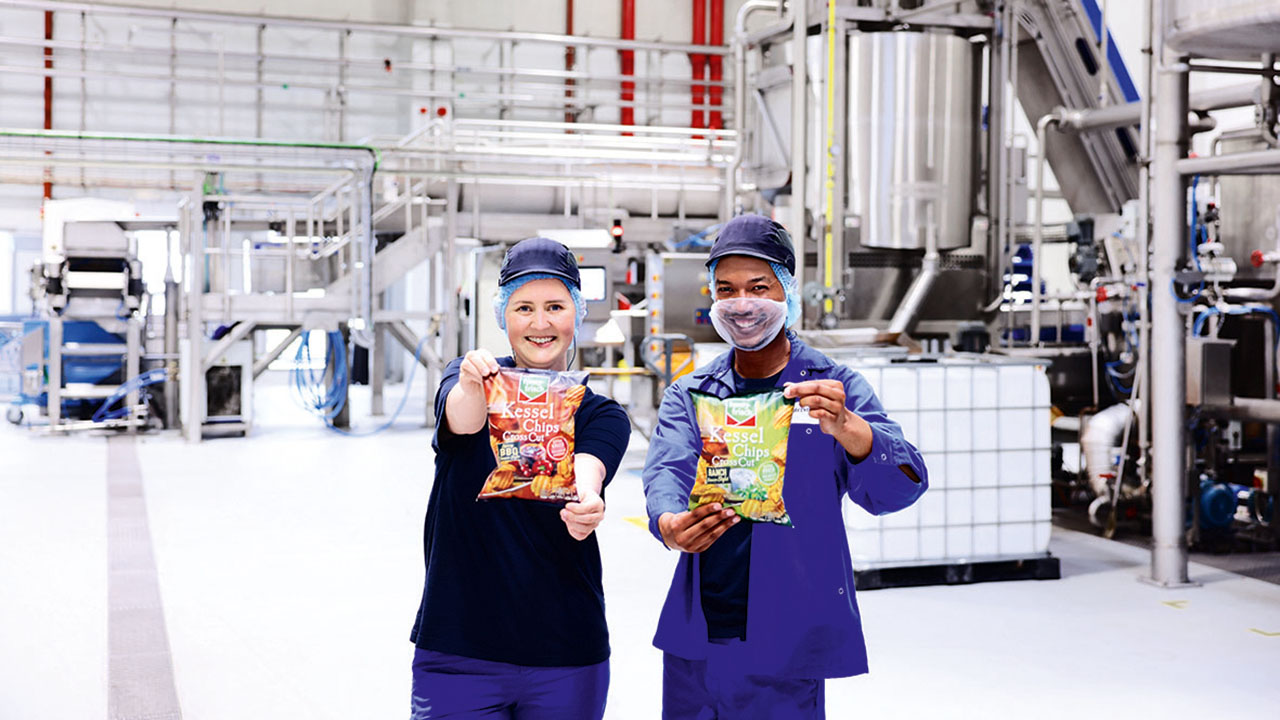 Lächelnde Mitarbeitende in Schutzkleidung halten eine Tüte Kesselchips in die Kamera