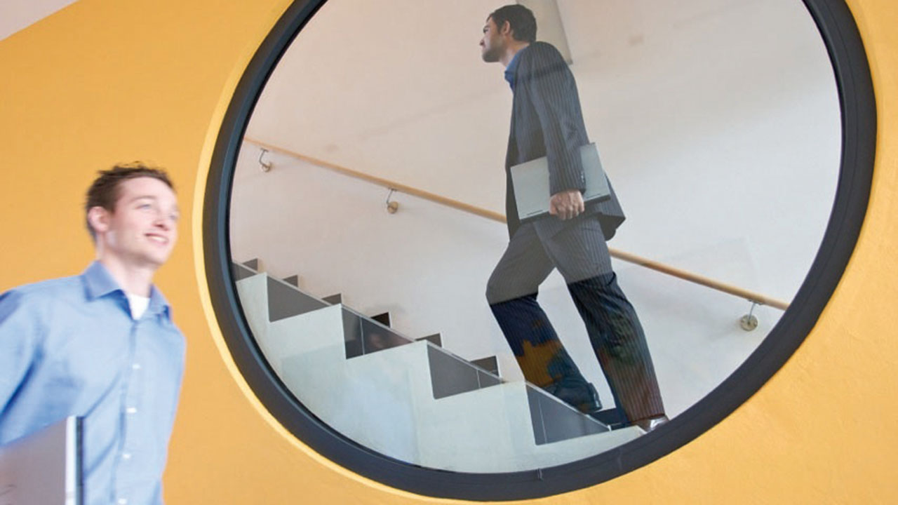 Junge lächelnde Person geht an einem kreisförmigen Fenster vorbei, durch das man eine Person im Anzug mit Laptop die Treppe hochgehen sieht