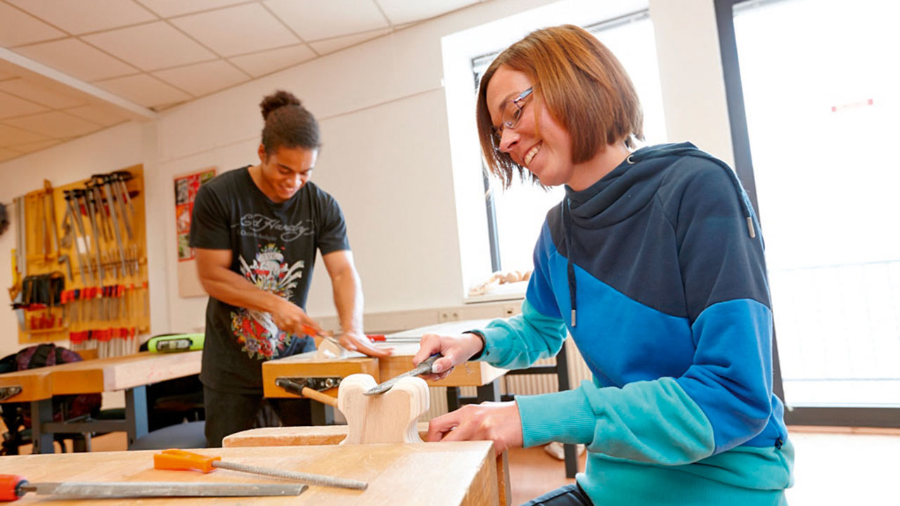 Zwei junge lächelnde Personen bearbeiten Holzstücke an einer Werkbank