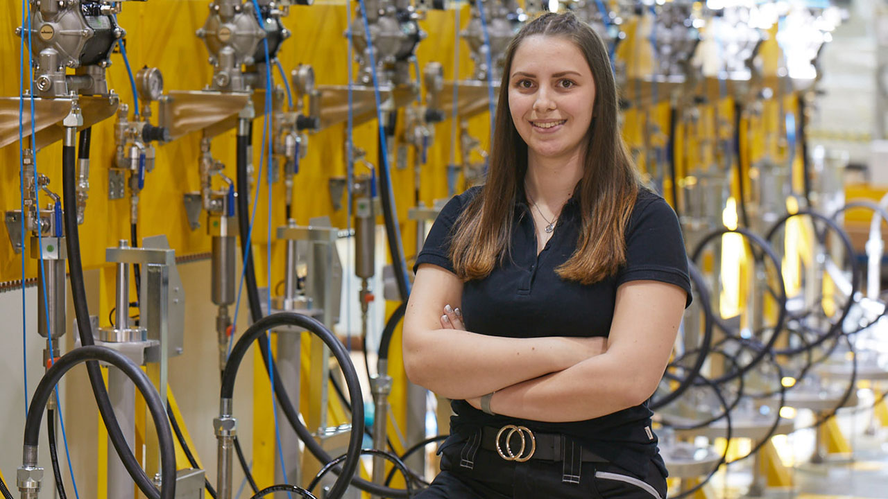 Eine Person mit langen Haaren steht in einer Werkhalle, trägt dunkle Kleidung und verschränkt die Arme. Im Hintergrund sind viele gelbe Maschinen und Schläuche zu sehen.