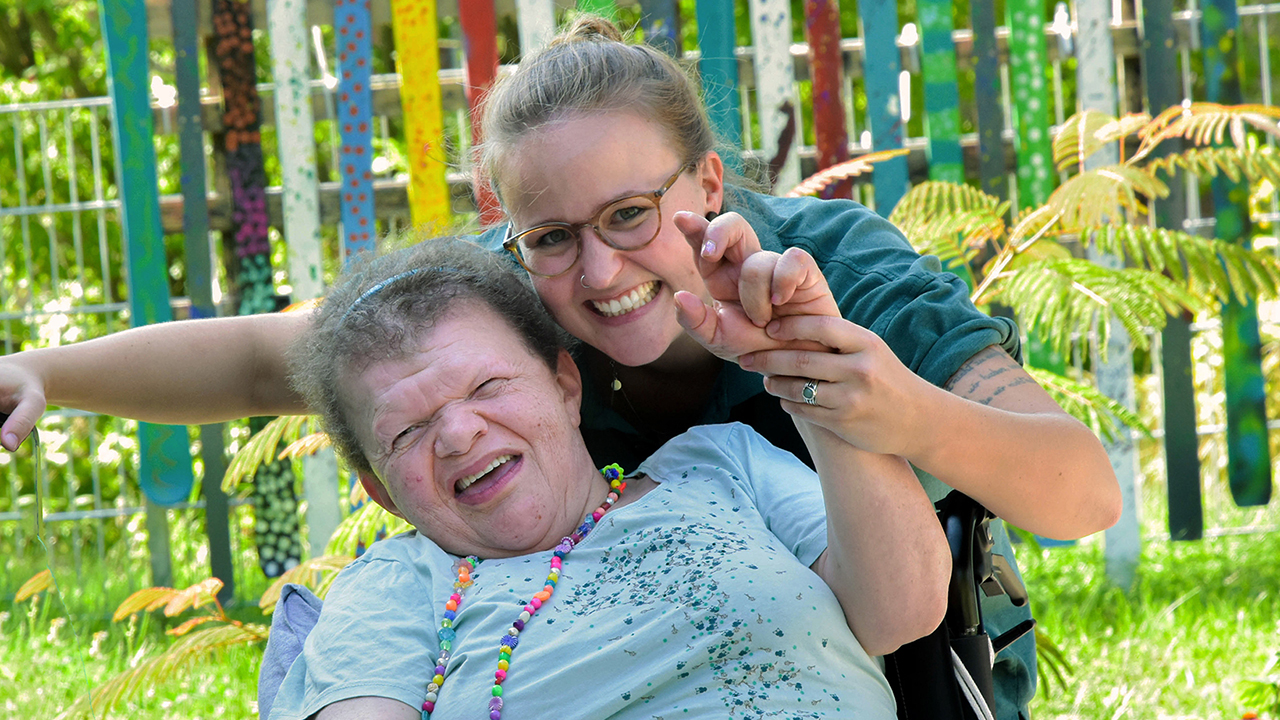 Zwei Personen lächeln in die Kamera, die hintere Person hält eine Hand der vorderen. 