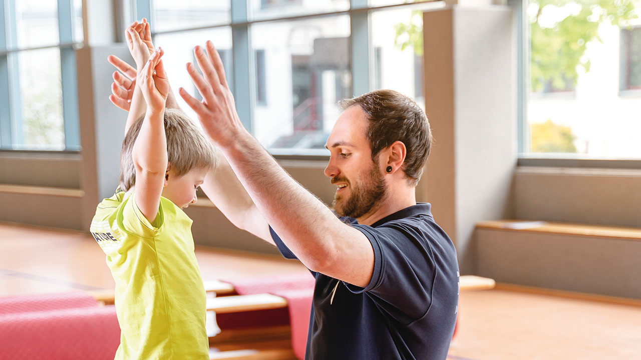 Ein Kind und eine erwachsene Person spielen zusammen. Das Kind kniet auf einem grünen Ball.