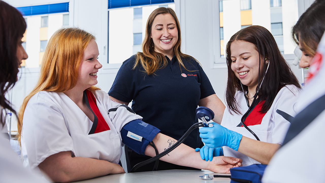 4 zukünftige Pflegefachkräfte beim Erlernen der Blutdruckkontrolle. Eine Lehrkraft begleitet die Übung.
